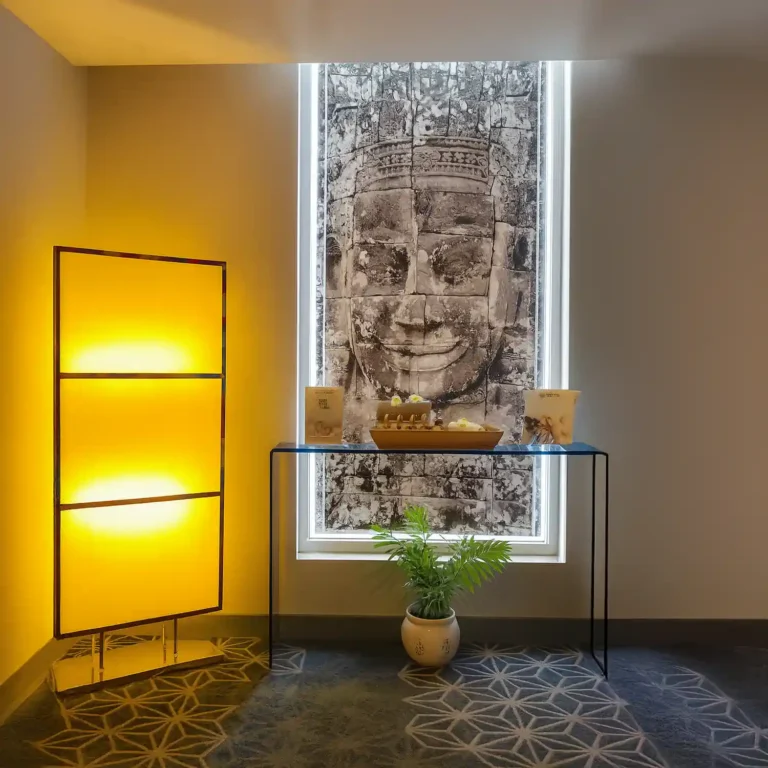 Decorative wellness corner at Tattva Wellness Spa, Four Points by Sheraton Chennai, featuring a serene Buddha wall art, soft yellow lighting, a minimalist table with spa oils and candles, and a potted plant for a calming effect.