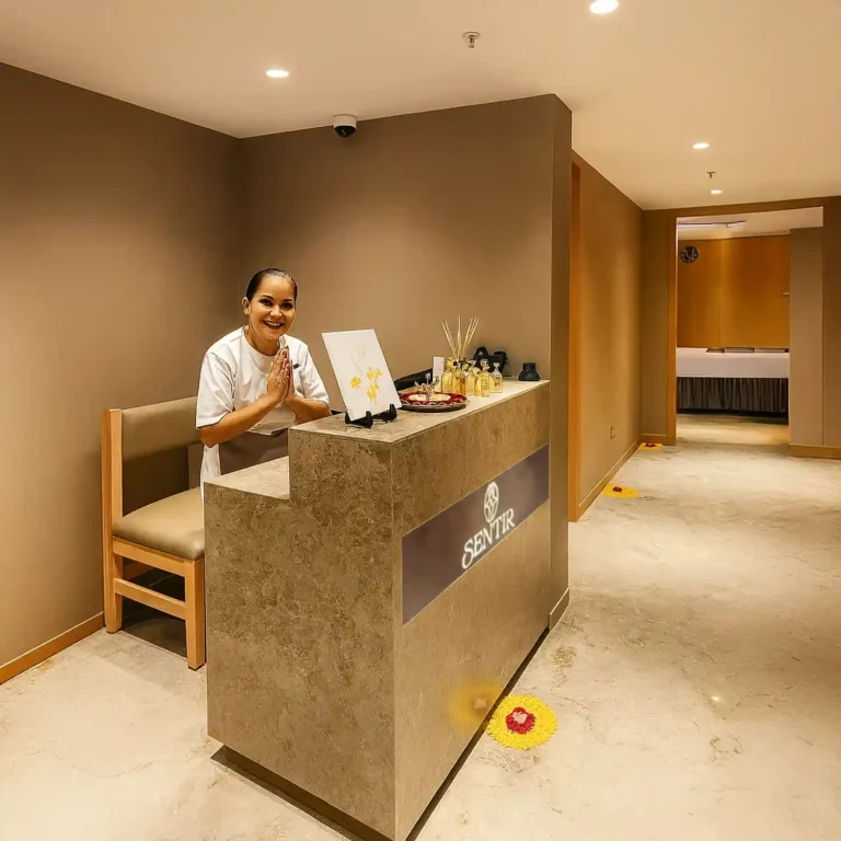 Warm reception area at Tattva Wellness Spa, Courtyard by Marriott Colva Goa, featuring a smiling healer greeting guests with folded hands beside a counter decorated with essential oils and flowers.