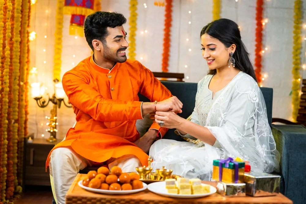 A man and woman celebrate Raksha Bandhan; the woman ties a rakhi on the man's wrist. Sweets, gifts, and festive decorations—including the best Rakhi gift for sibling—are beautifully arranged on the table in front of them.
