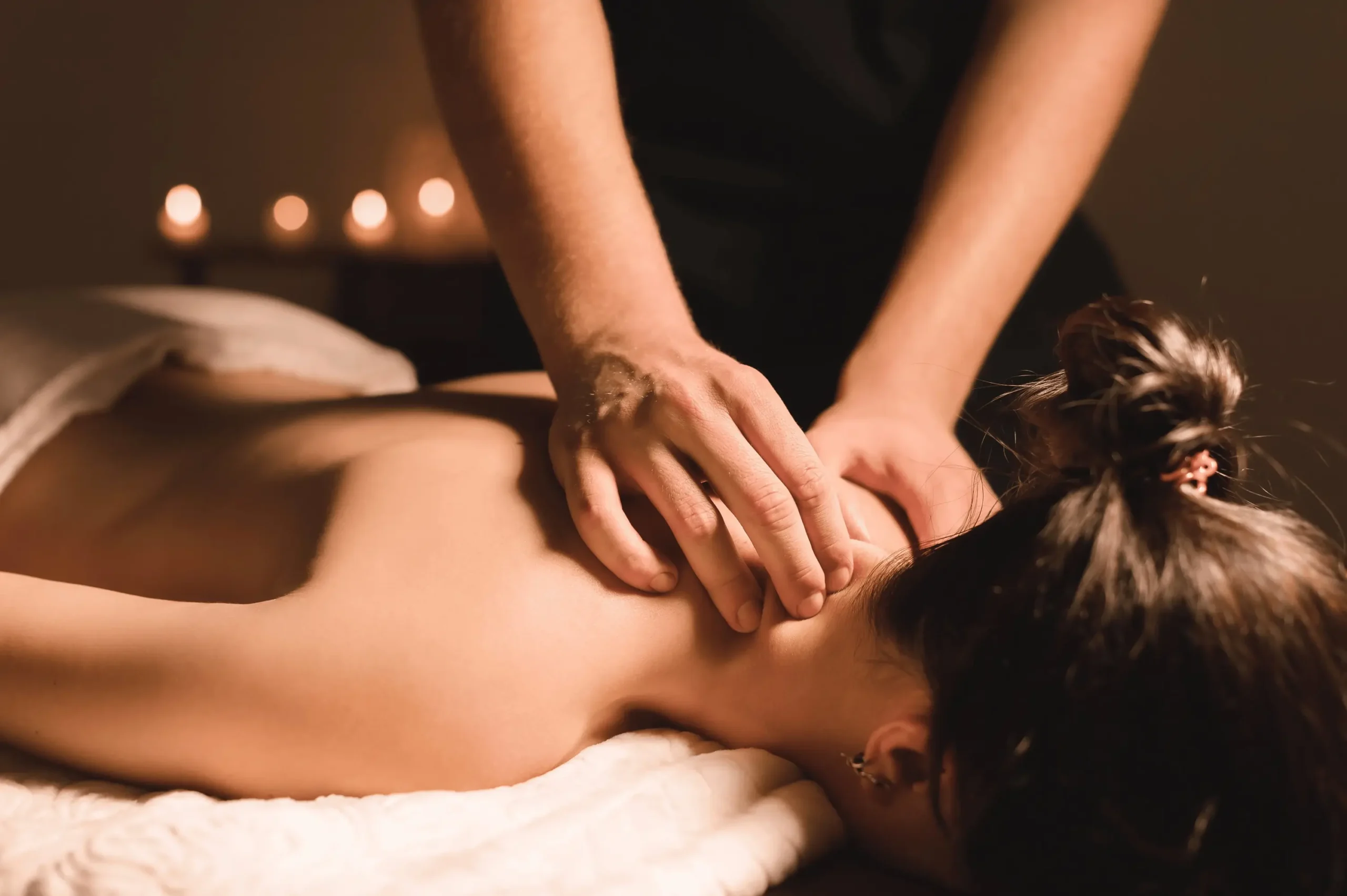 A woman receives a relaxing Swedish massage in a serene spa setting; a healer applies gentle pressure along her shoulders and upper back. The room is softly lit with candles, creating a calming ambiance ideal for therapeutic relaxation.