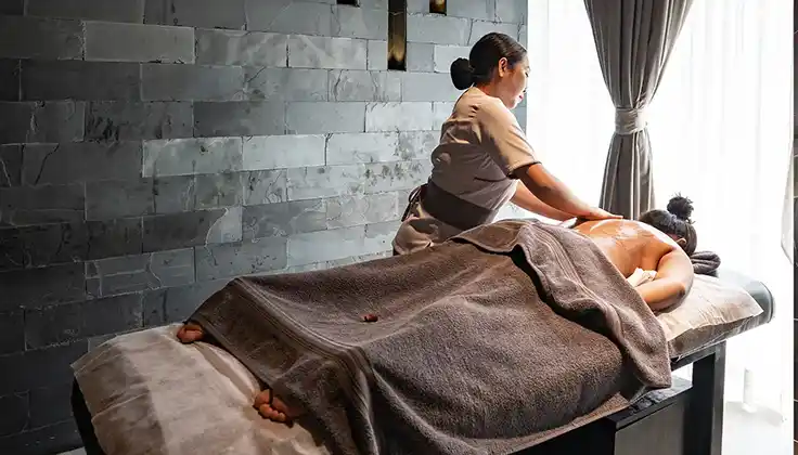 A massage therapist gives the best Swedish massage in Lonavala to a person lying face down on a massage table, covered with a towel, in a softly lit spa room.
