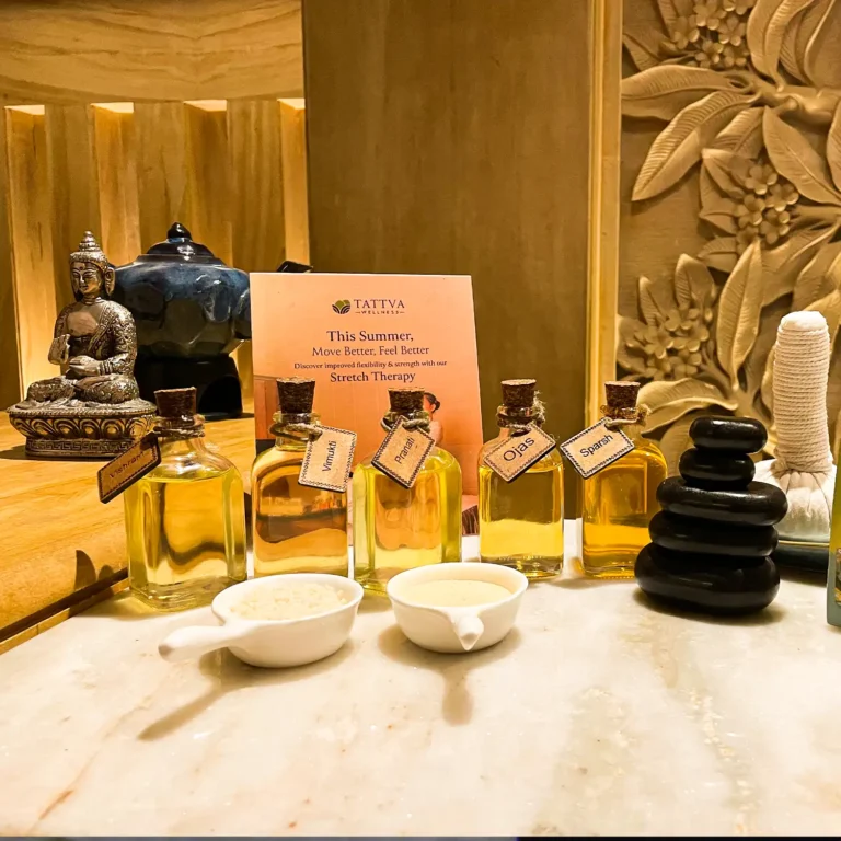 A close-up display of spa therapy essentials arranged on a marble counter, including glass bottles filled with massage oils, small white bowls containing herbal ingredients, stacked black hot stones, and a rolled herbal compress, set against a warm golden spa interior.