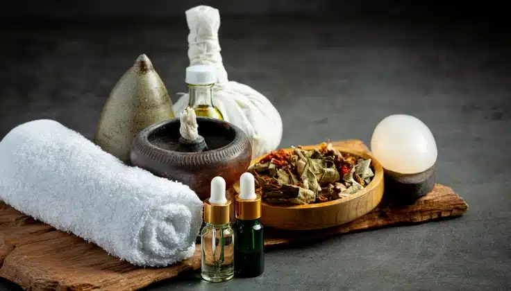 Ayurvedic spa setup with herbal compresses, massage oils, dried herbs and a rolled white towel.