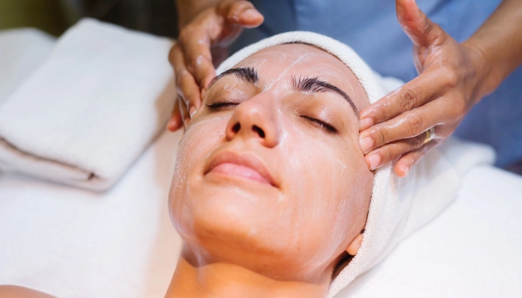 Therapist gently massaging a woman's face while applying facial cream during a spa treatment.